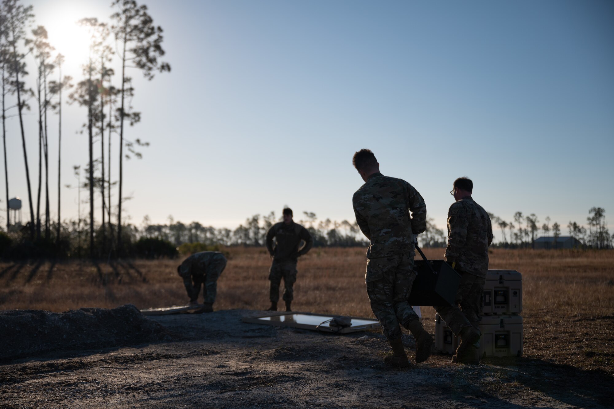 Two Airmen carry a box towards two more Airman who are setting up an electrical system in a field.