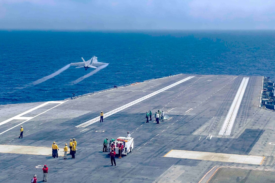 Sailors stand on the deck of a ship as a jet flies above.