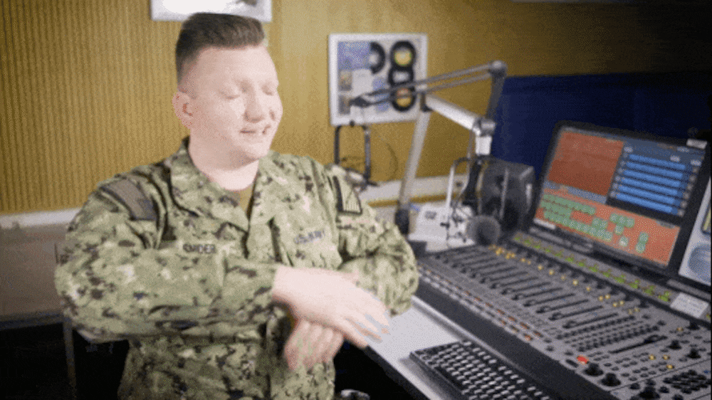 A service member, with his hands up, apart and palms facing in, is moving his hands close together to demonstrate the hand signals and movements for communicating to a guest to lean in closer to the microphone.