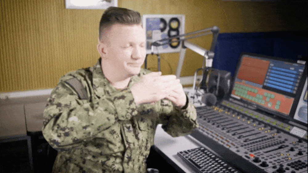 A service member, with his hands up, close together and palms facing in, is moving his hands apart to demonstrate the hand signals and movements for communicating to a guest to lean away from the microphone.