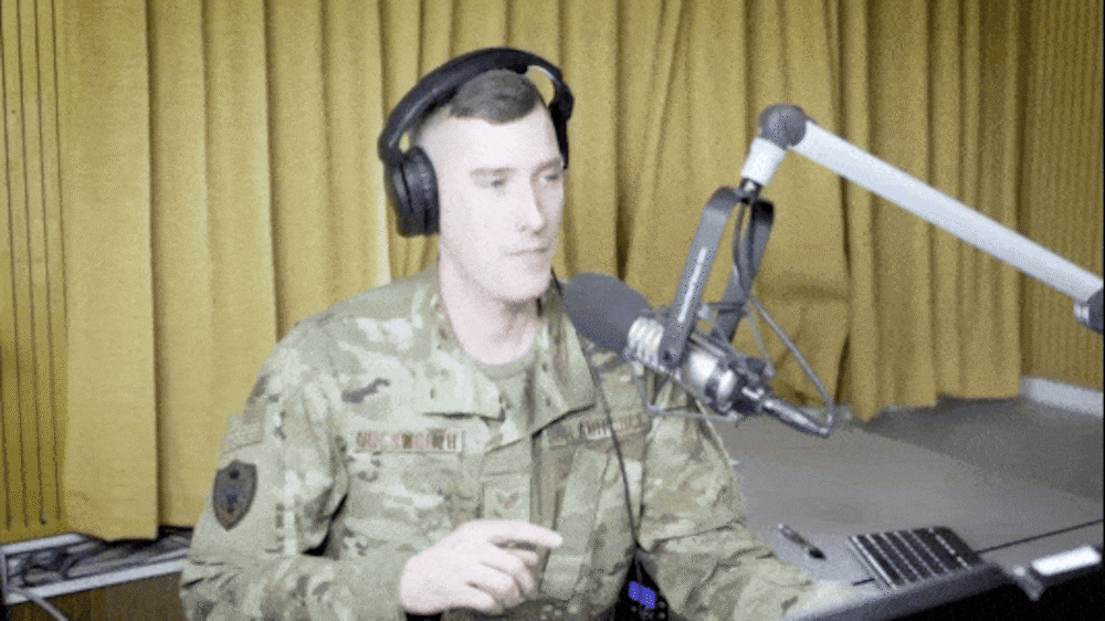 A service member holds up his index finger in the air, next to his head. He circles his finger in the air to communicate to a guest to finish their sentence.