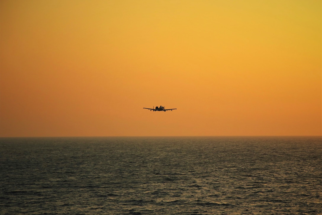 An aircraft flies over a body of water under an orangish sky.