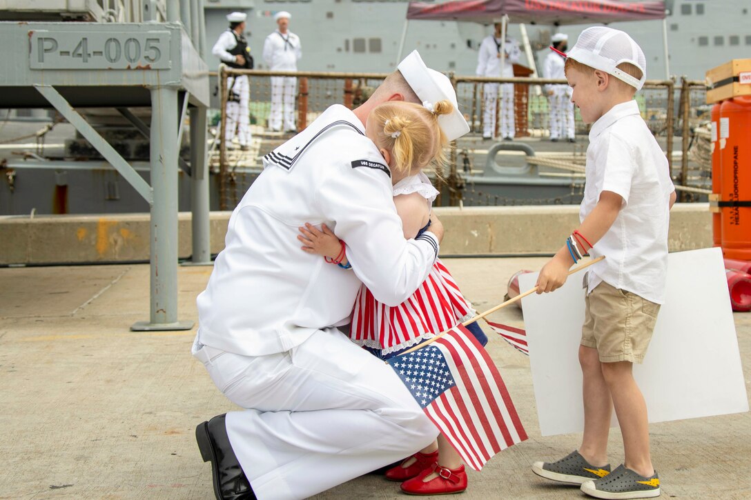 A sailor kneels to hug a child as another child standing nearby watches while carrying an American flag.