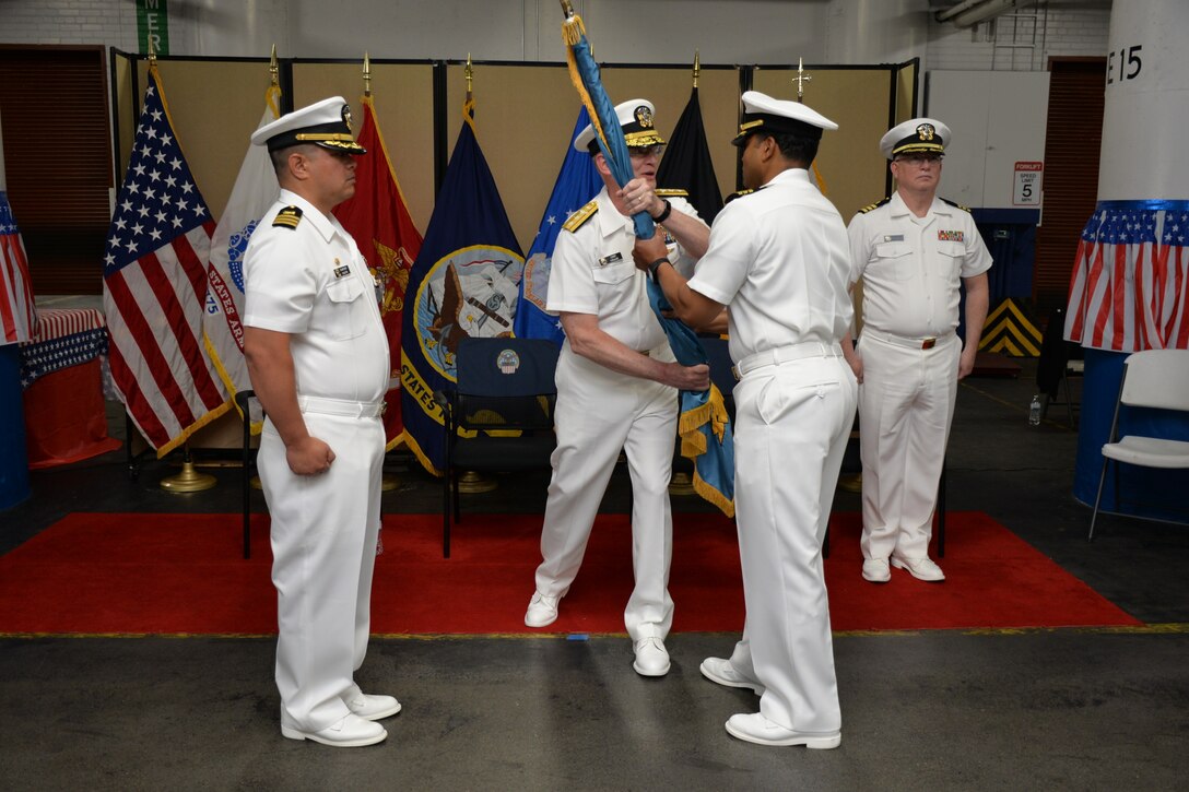 Photo is of Navy officers dressed in white, with one officer passing a flag to another