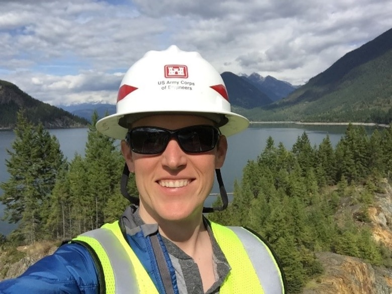 Photo of Caucasian man wearing a hard hat and safety vest taking a selfie with mountains in the background.