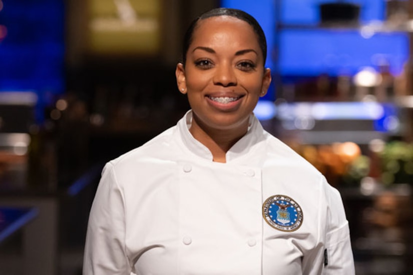 A woman in a chef's uniform is photographed in a kitchen.