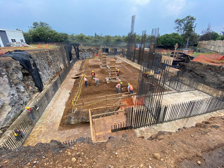 Several construction workers can be seen working on the foundation for a building with trees and a sky in the background.