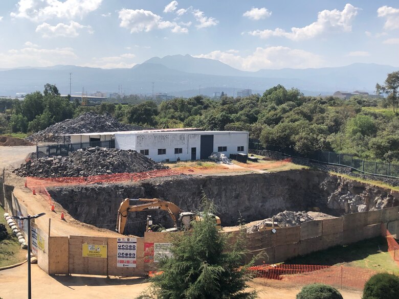 A large hole is dug in the ground, surrounded by safety fencing, with construction equipment in the foreground. Trees, mountains, and the sky can be seen in the background.