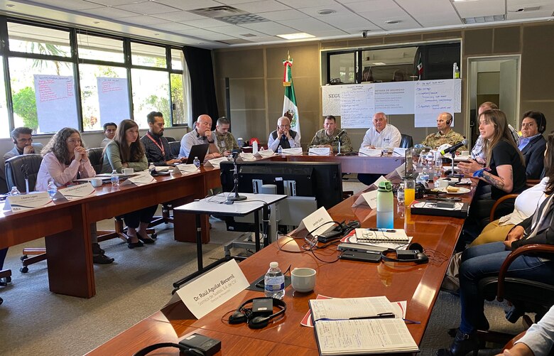 Many people sit around a conference table in the shape of a U with the Mexican flag in the background.