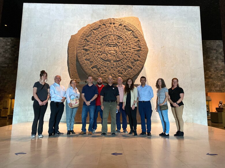 Eleven people stand in front of a large archeological plaque in the foyer of a museum.