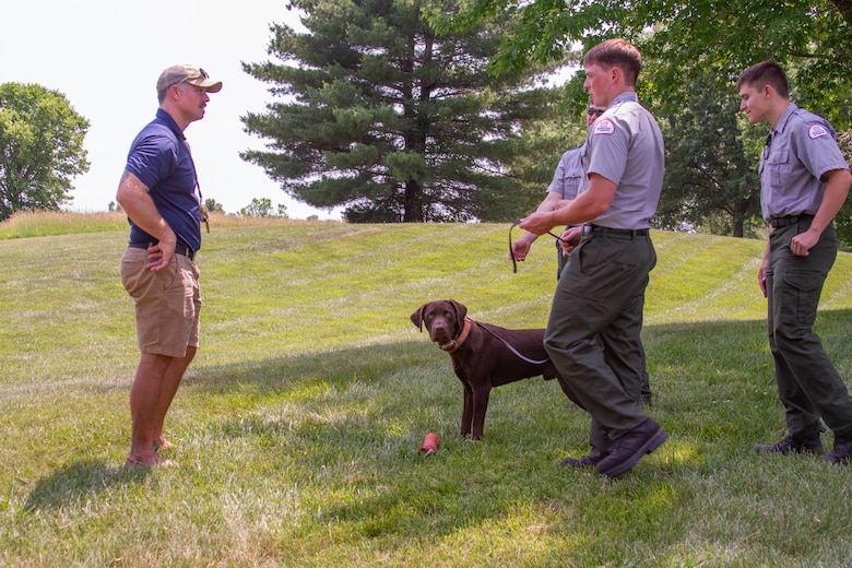 Park rangers speak to a man with a dog in the grass with trees in the background.