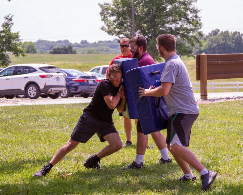 Four people engage in self defense training with grass in the foreground, cars and trees in the background.