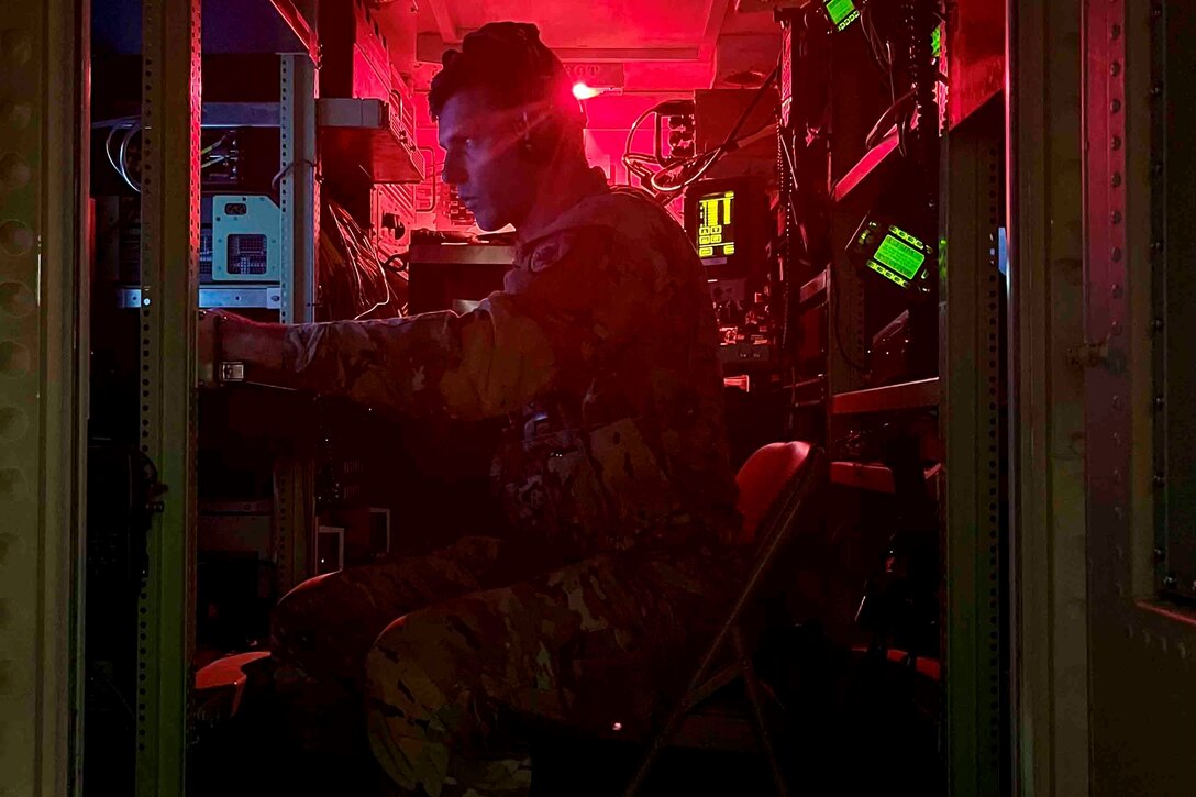 A soldier illuminated by red light sits on a chair between shelves while working on equipment.
