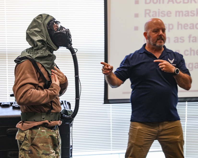 Two men stand in the foreground. On the left, an Airmen demonstrates how to wear the Joint Service Aircrew Mask for Strategic Aircraft (JSAM-SA) and the trainer stands in civilian clothes on right during an instructional briefing.