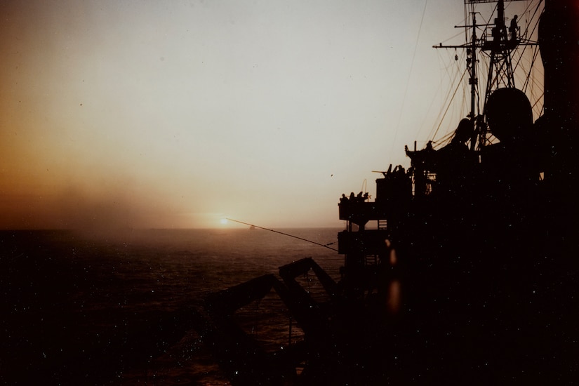 A sun rises over the ocean. To the right, parts of a ship are seen in silhouette.