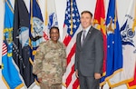 A dark skinned woman in a camouflaged uniform stands next to a light skinned man in a dark grey suit with graying hair in front of 8 military flags.