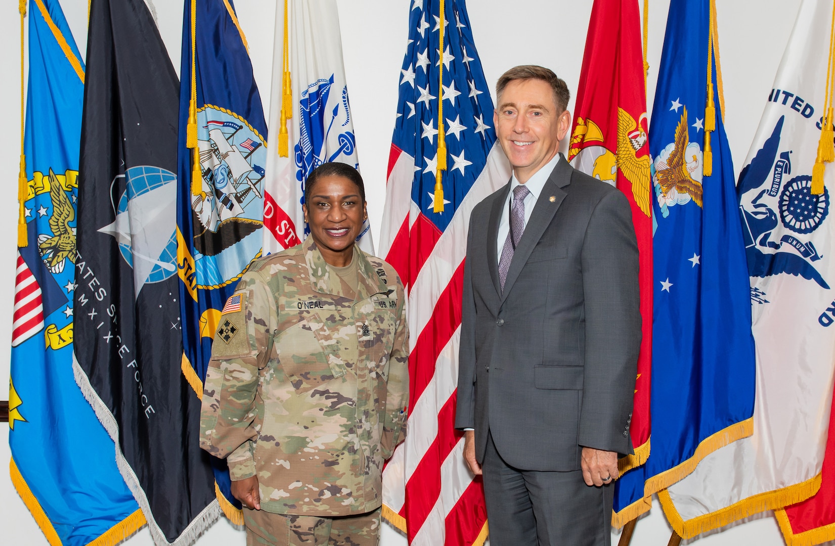 A dark skinned woman in a camouflaged uniform stands next to a light skinned man in a dark grey suit with graying hair in front of 8 military flags.