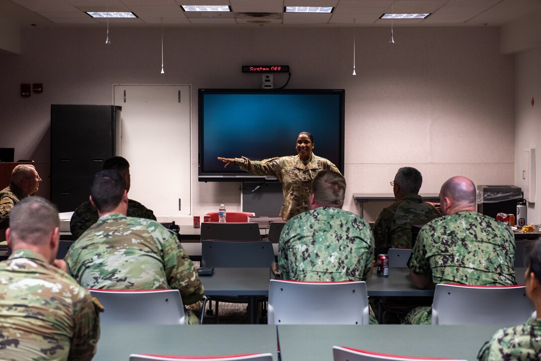 A dark skinned woman in a camouflaged uniform speaks to other military members in a darkened room.
