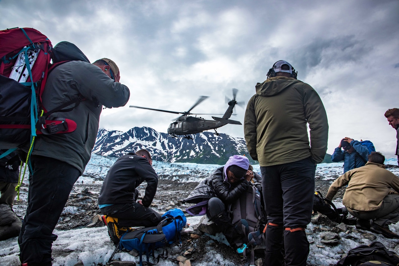 Crew members spread pack up and prepare to leave a glacier with a helicopter and mountain peaks in the distance.