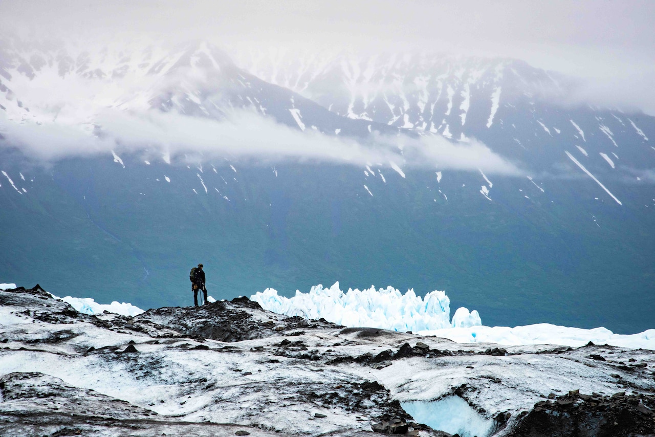 A soldier looks out toward mountains during a search along a ridge on a glacier.