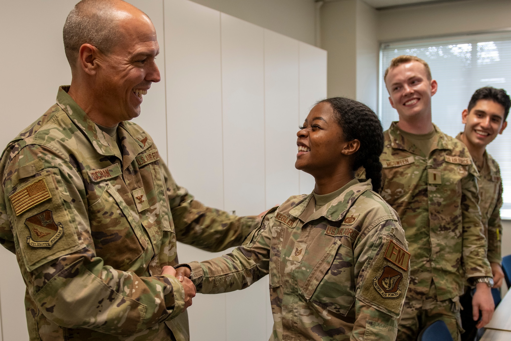 A tall male commander shakes hands with a shorter female soldier, they're both grinning