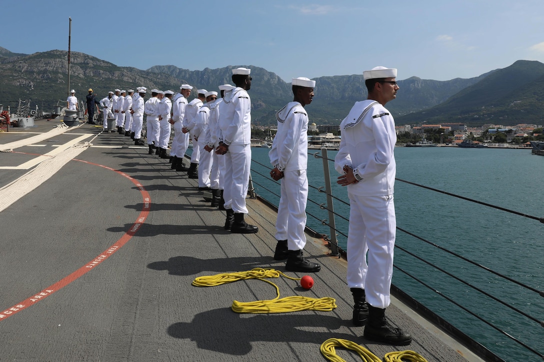Sailors stand at parade rest while a ship pulls into port.