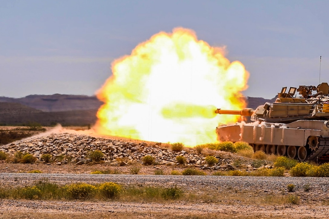 A blast emits from the front of a military tank in a desert area.