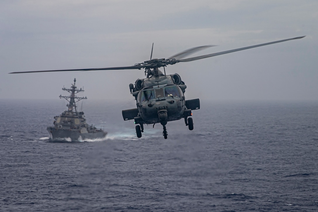 A military helicopter flies over open water as a Navy ship sails in the background.