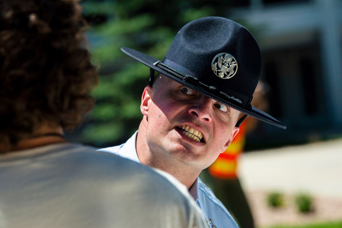 An airman grits his teeth and opens his eyes wide while staring at a basic cadet with greenery in a blurred background.