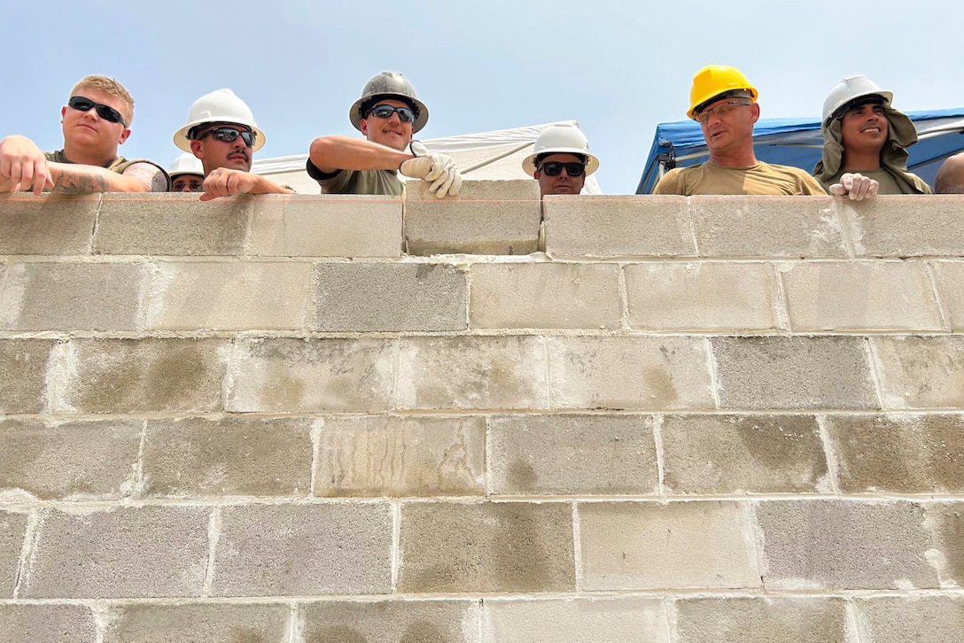 Airmen wearing hard hats while standing along a cement wall place a block on that wall.