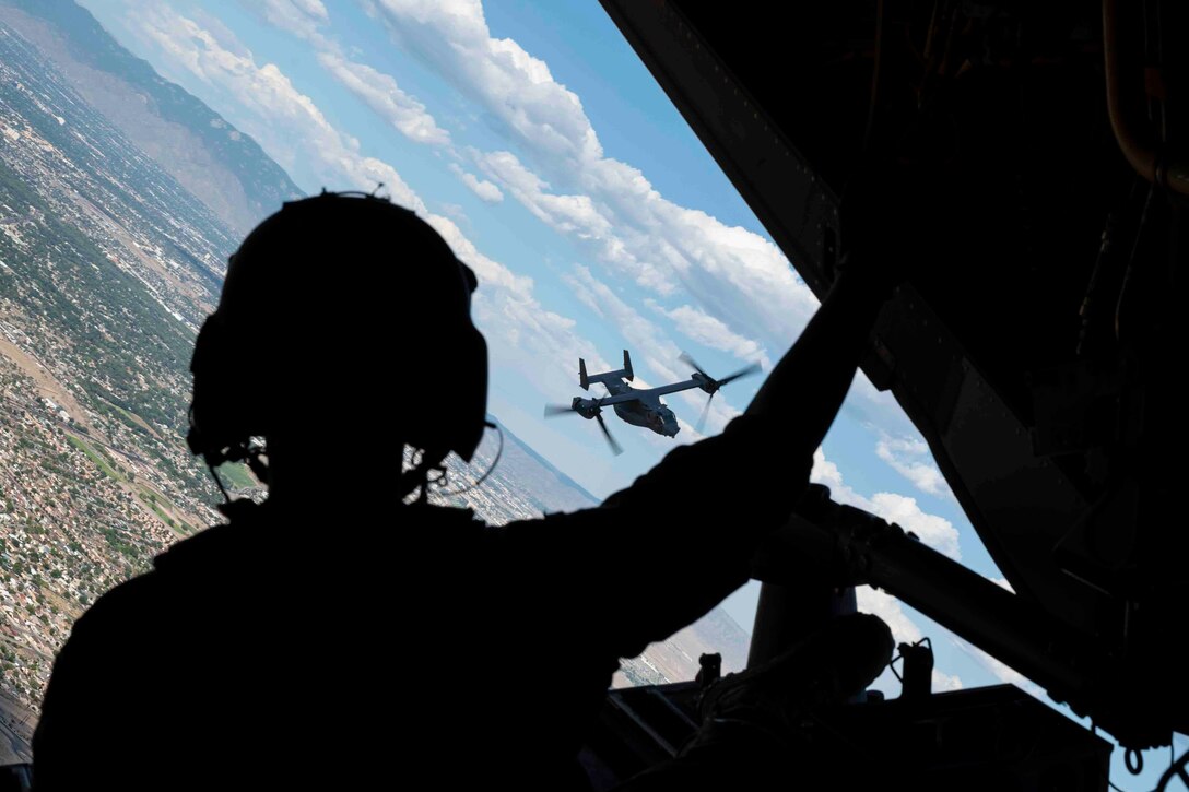 An airman looks out from the back of an airborne aircraft as another aircraft flies over a city.