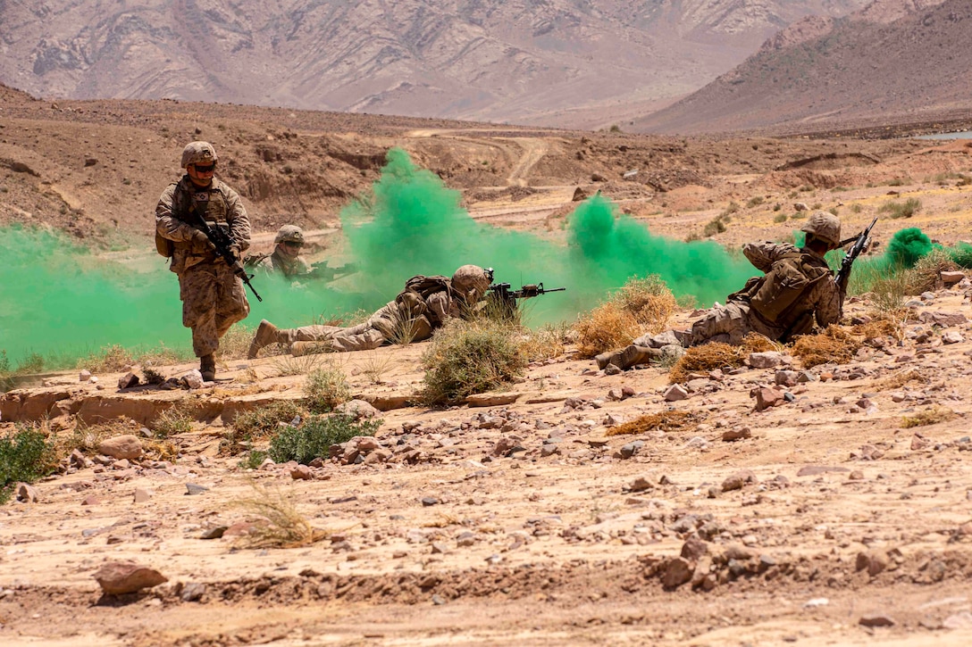 Four Marines lie, kneel and stand in desert terrain as green smoke moves between them.