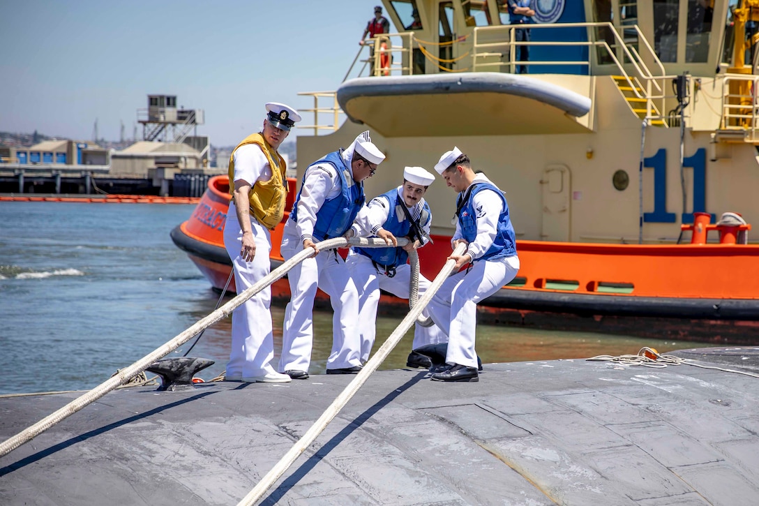 Sailors pull a rope while standing on top of a submarine in front of a boat.