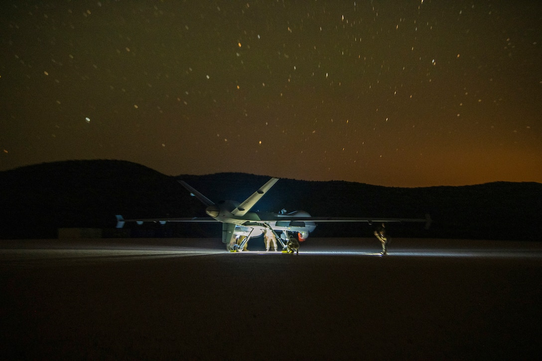 Airmen work on an unmanned aircraft parked on a dirt road illuminated by a spotlight under an orangish starry night sky.