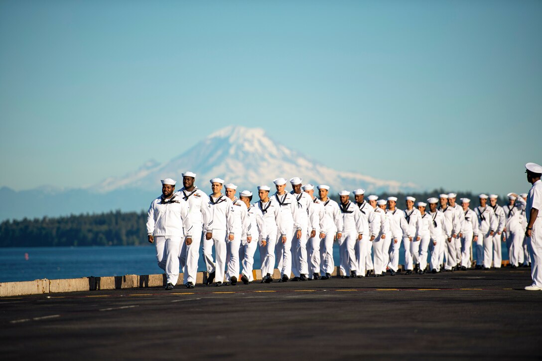 Sailors walk in formation aboard a ship with a body of water, greenery and a mountain in the background.