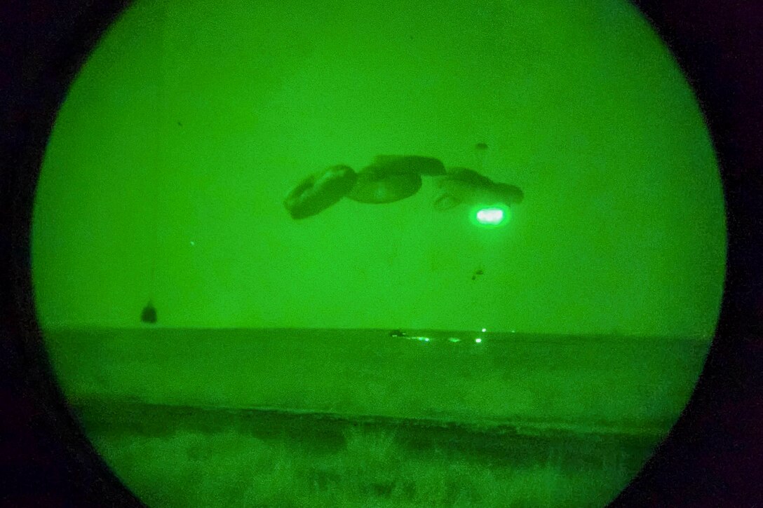 Soldiers shinning flashlights descend with parachutes into a field as seen through night vision goggles.