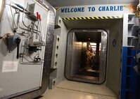 An open vault door frames three Air Force personnel standing inside a missile launch control center. "Welcome To Charlie" is painted above the entrance.