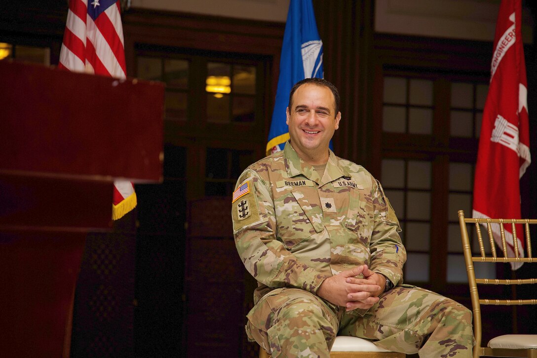Photo shows military officer sitting and smiling while listening to remarks