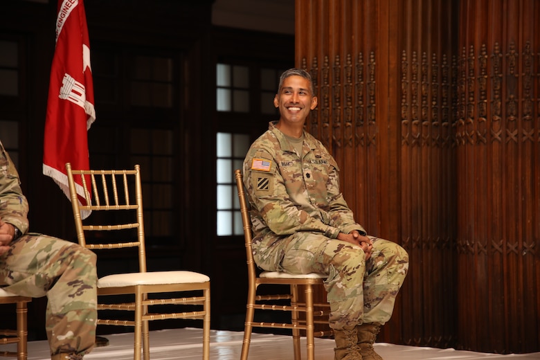 Photo shows a military officer sitting and smiling while hearing remarks during a ceremony