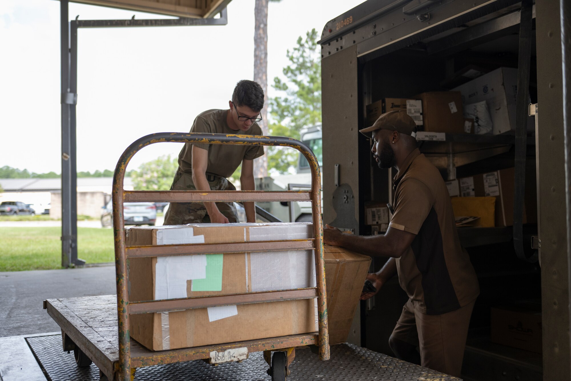 A photo of an airmen placing boxes on a trolley while a mail carrier worker is handing them up on the mail deck.