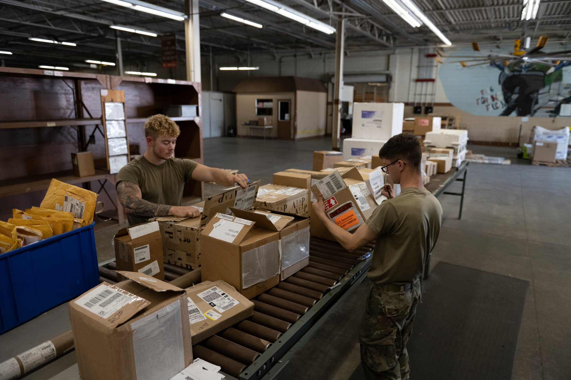 A photo of two people in a ware house with boxes on a conveyer belt in front of them.