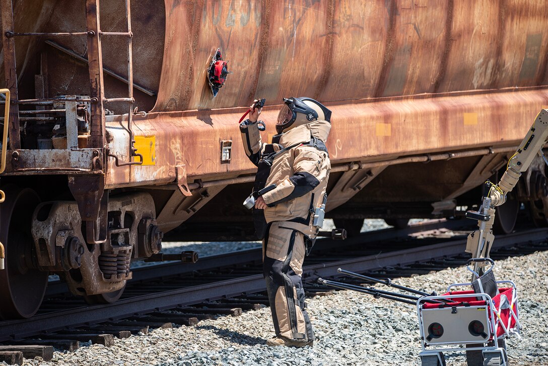 An airman in a protective suit examines a simulated explosive device on a rail car during training.