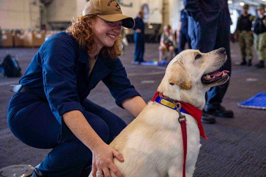 A sailor squats next to a dog while petting it.