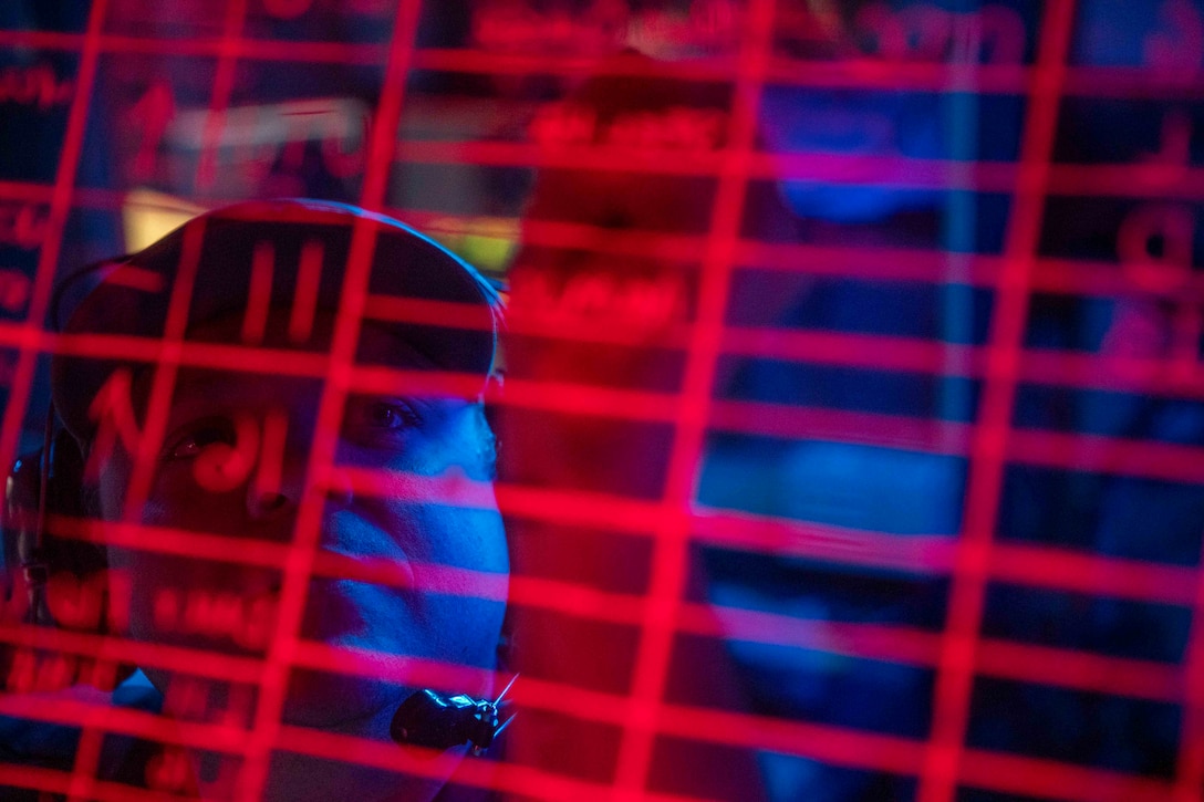 A closeup of the reflection of a sailor wearing a headset writing in the lines on a blackboard illuminated by red light.