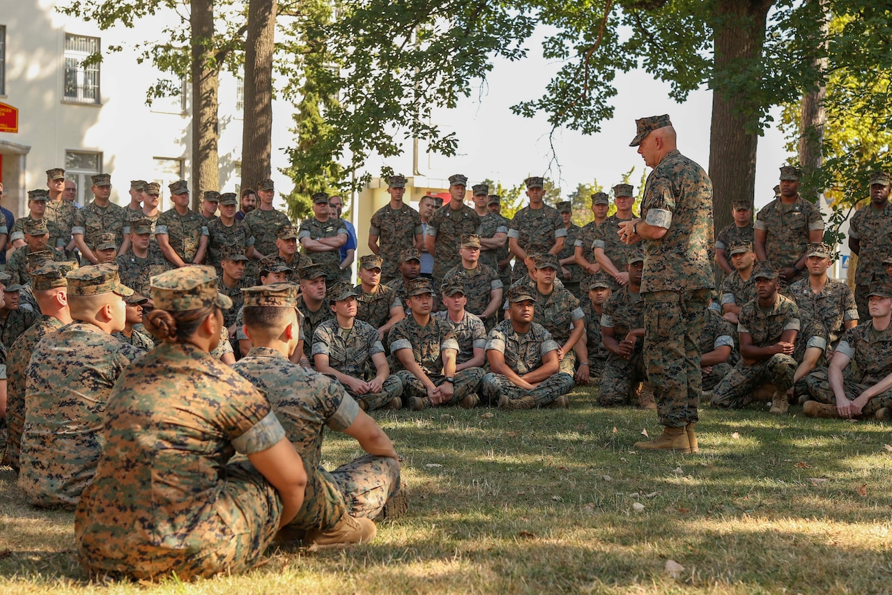 A man in uniform speaks to a group of Marines and sailors.