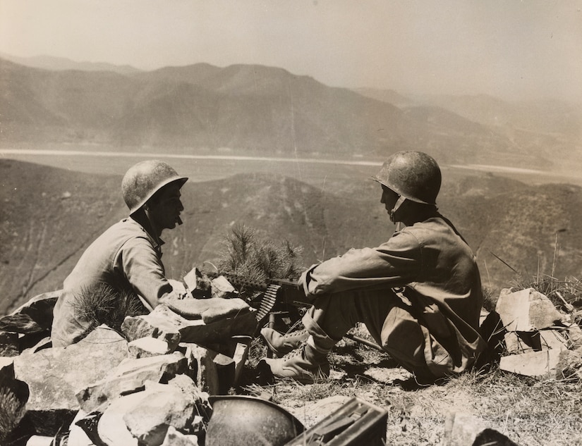 Two men in battle gear sit on the ground surrounded by equipment, looking out over vast terrain.