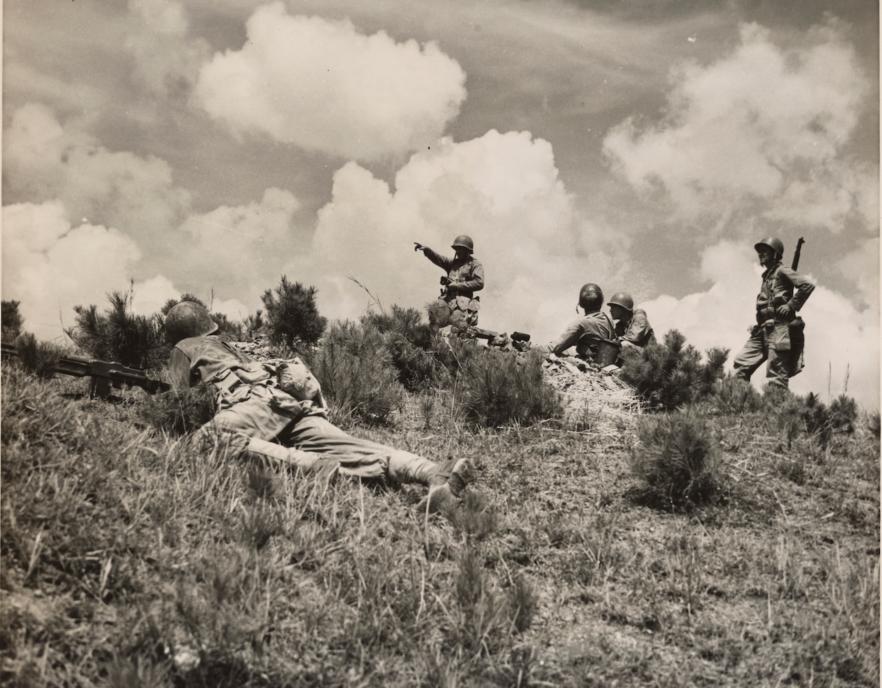 A soldier with a machine gun lies on a hill as other soldiers stand and squat further away from him.
