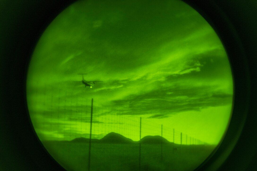 An aircraft flies over mountains and a fenced field as seen through night-vision lens.