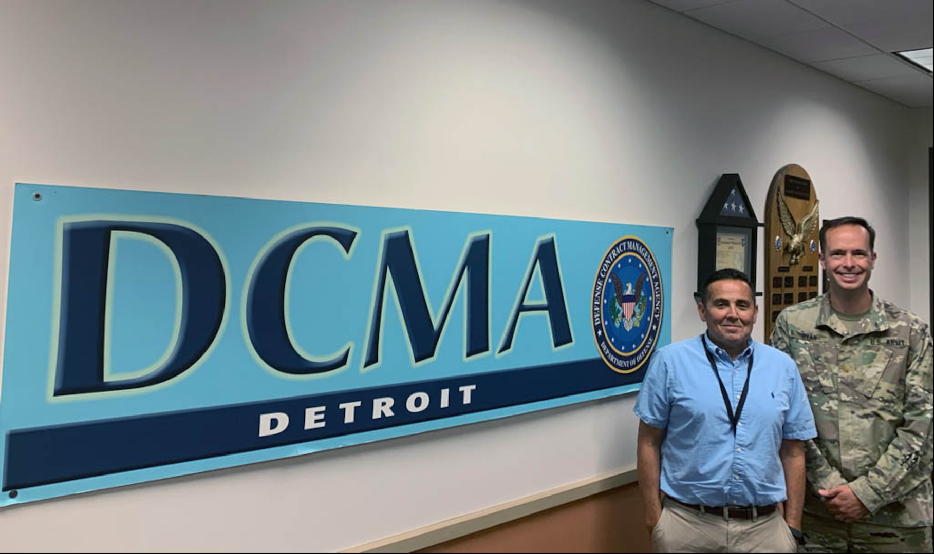 One man wearing a blue shirt and khaki pants and another man in his Army uniform stand near the DCMA Detroit sign