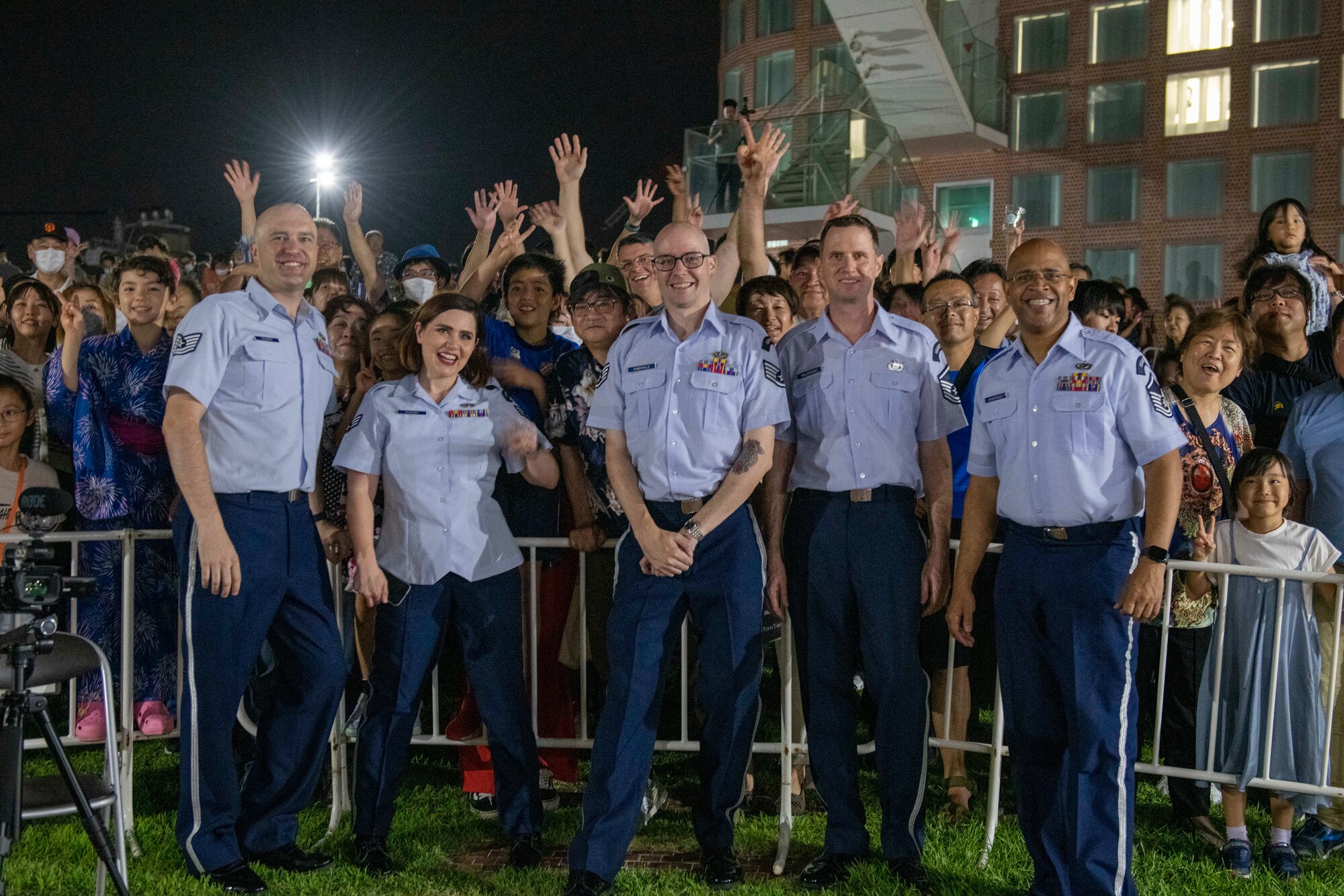 Four military band members pose for a photo with dozens of fans in the background waving